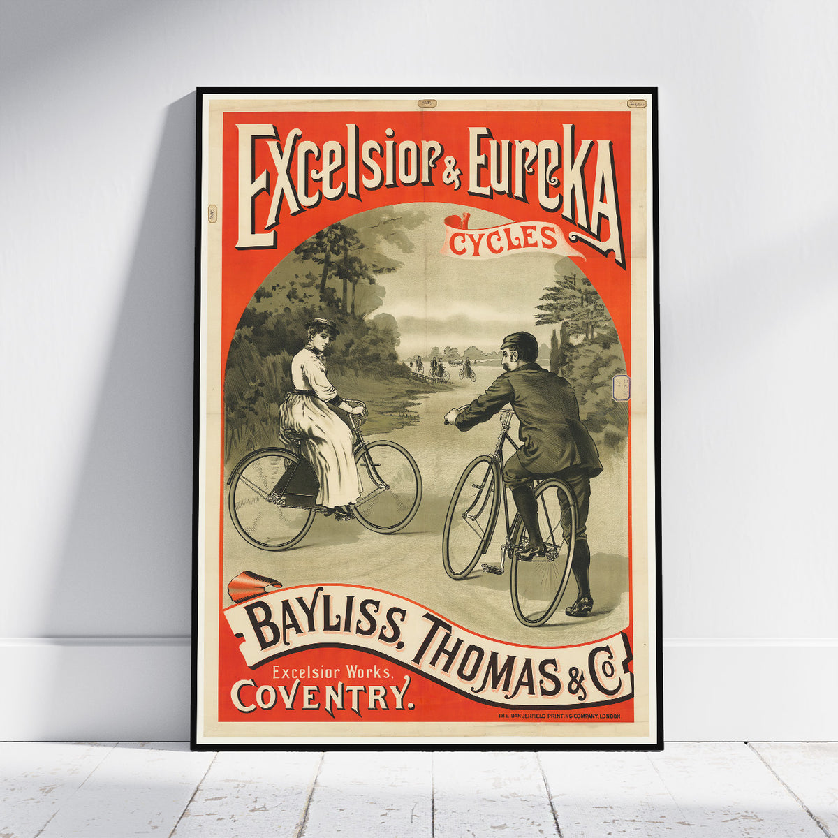A framed vintage cycling advertisement poster leaning against a white wall on a light wood floor. The poster features an orange-red border, sepia-toned illustration of two people riding bicycles, and large text reading 'Excelsior & Eureka Cycles' and 'Bayliss Thomas & Co.