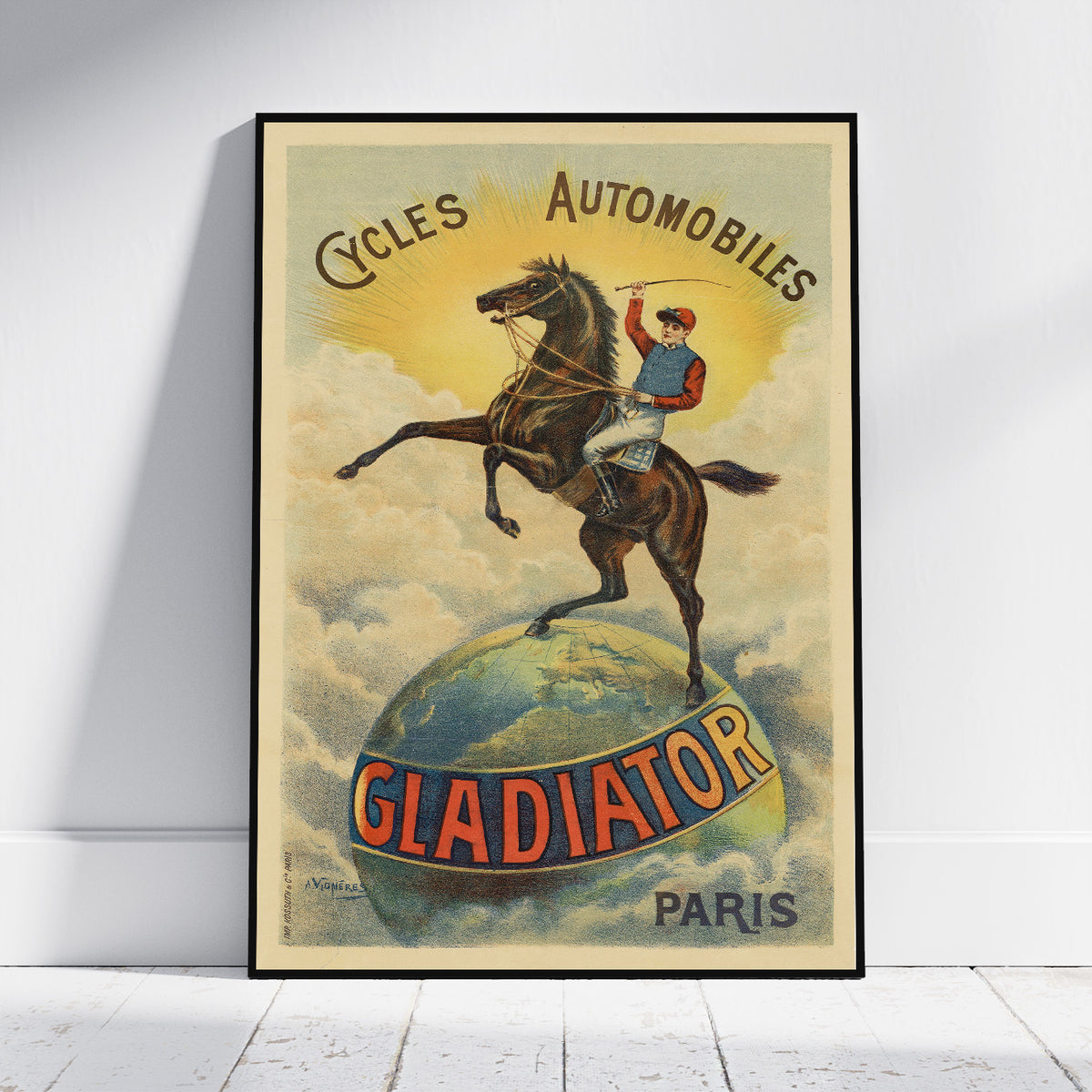 A framed vintage-style poster leaning against a white wall on a light wood floor. The poster shows a rider on a rearing horse standing on a globe with the text 'Cycles Automobiles' and 'GLADIATOR PARIS'.