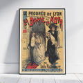 A framed vintage theatrical poster leaning against a white wall on a light wood floor. The poster features illustrated figures in formal 19th-century clothing with large red title text reading 'La Dame en Noir' and additional French text at the top and bottom.