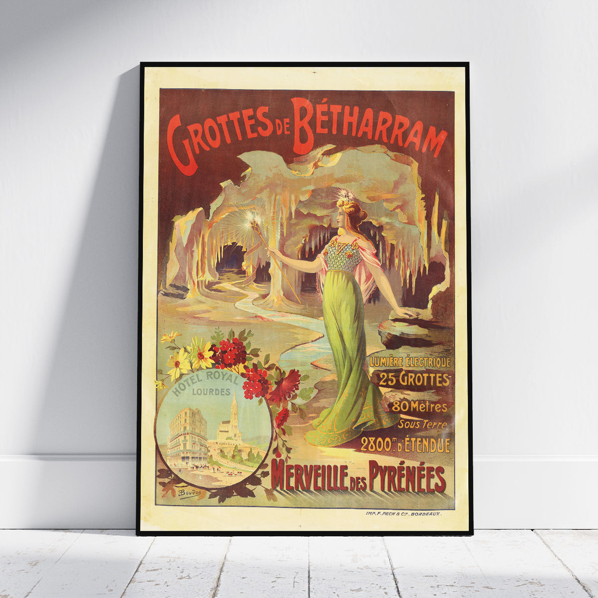 A framed vintage travel poster for 'Grottes de Bétharram' leaning against a white wall on a light wood floor. The poster features an illustration of a woman in a green dress in a cave, with red lettering and a cream border.