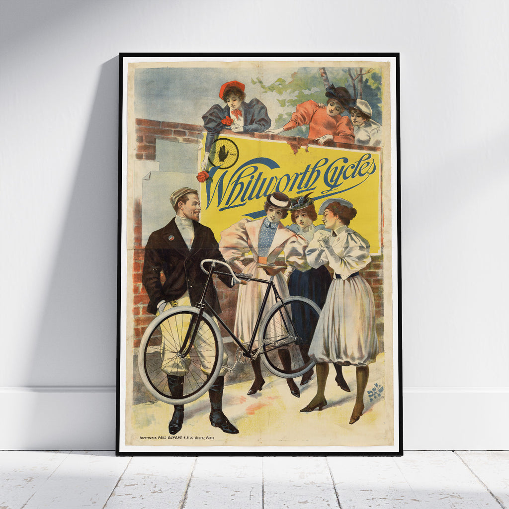 A framed vintage cycling poster leaning against a white wall on a light wood floor. The poster features an illustrated scene with several people around a bicycle and a large yellow sign reading 'Whitworth Cycles'.