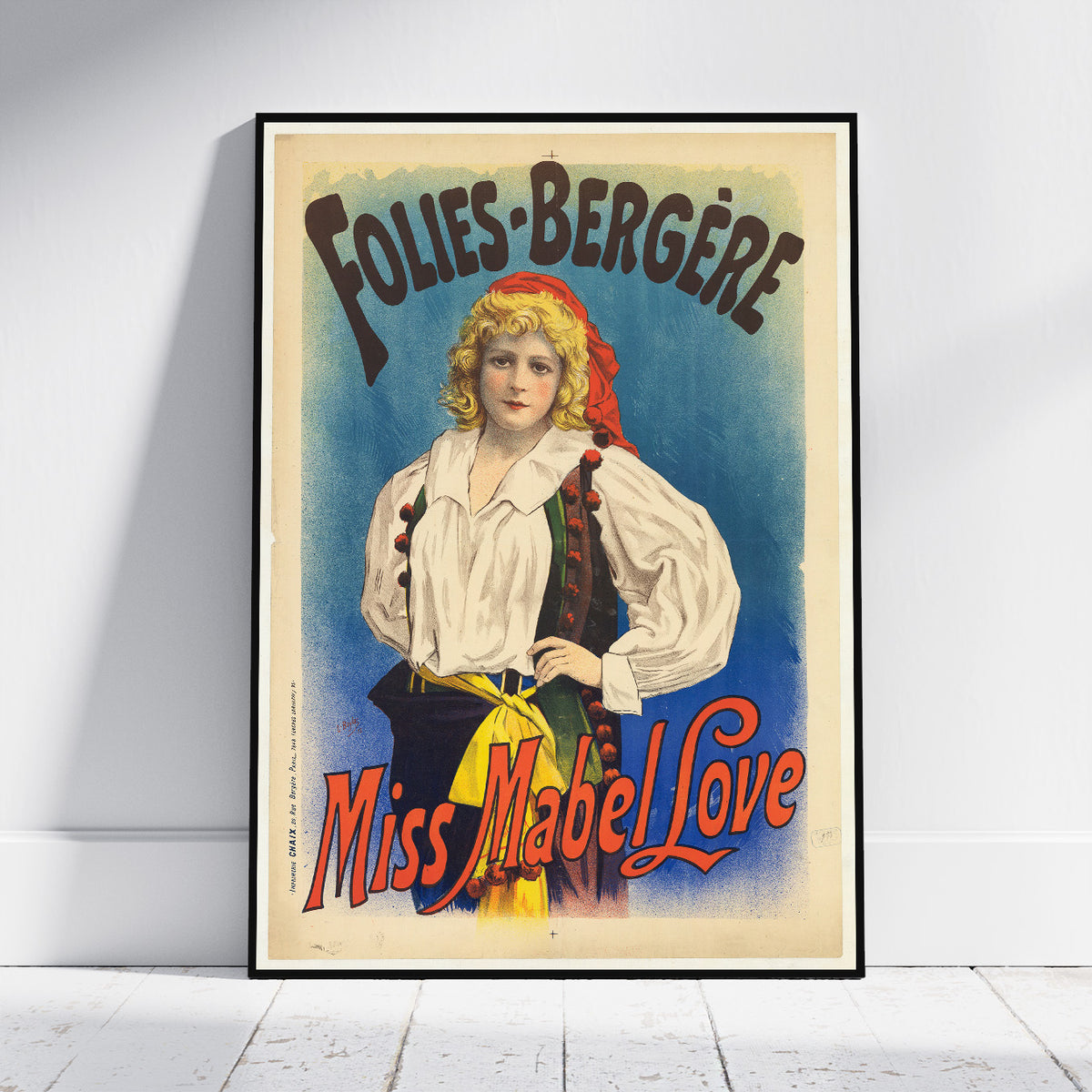 A framed vintage theatrical poster leaning against a white wall on a light wood floor. The poster features an illustrated woman in a white blouse and dark skirt with red and yellow accents, set against a blue background, with the text 'FOLIES-BERGÈRE' at the top and 'Miss Mabel Love' at the bottom.