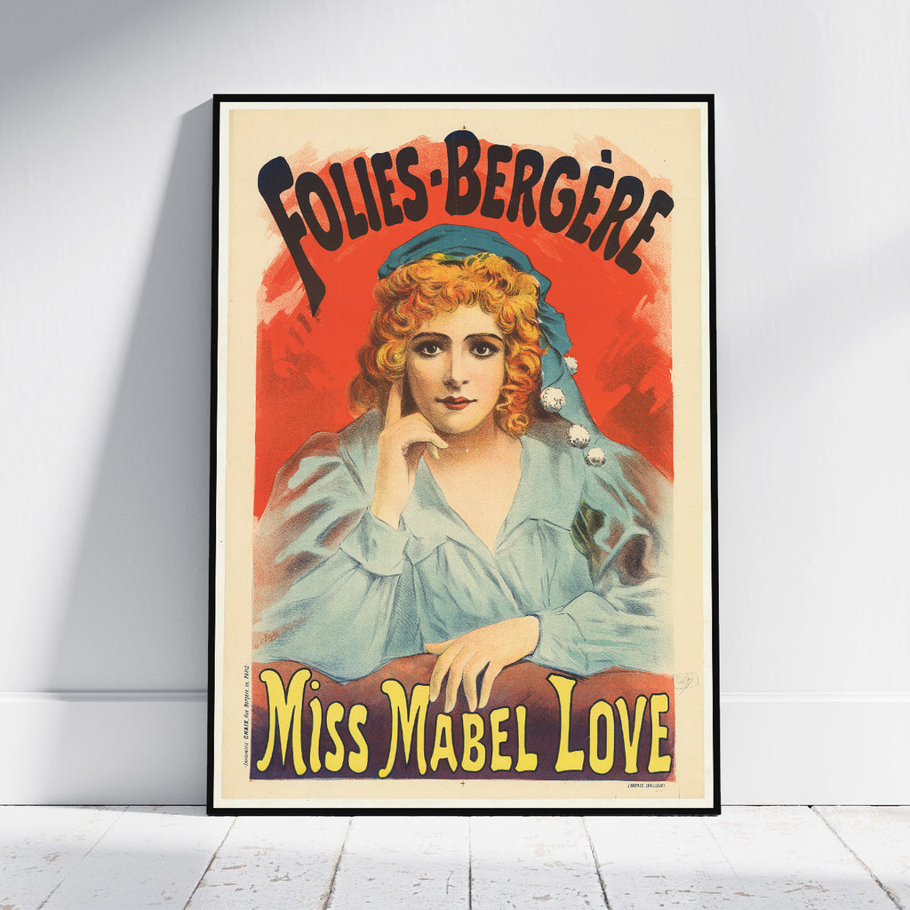 A framed vintage-style poster leaning against a white wall on a light wood floor. The poster features an illustrated woman with red hair and a blue head covering, with large text reading 'FOLIES-BERGÈRE' at the top and 'MISS MABEL LOVE' at the bottom.