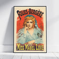 A framed vintage-style poster leaning against a white wall on a light wood floor. The poster features an illustrated woman with red hair and a blue head covering, with large text reading 'FOLIES-BERGÈRE' at the top and 'MISS MABEL LOVE' at the bottom.