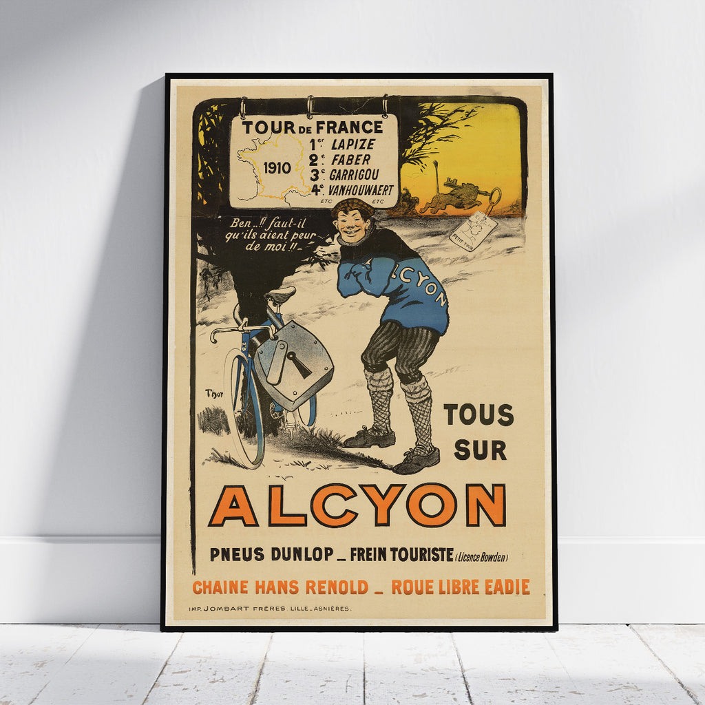 A framed vintage-style poster leaning against a white wall on a light floor. The poster features an illustrated cyclist and rider with the text 'TOUR DE FRANCE 1910' and large 'ALCYON' lettering, plus 'TOUS SUR'.