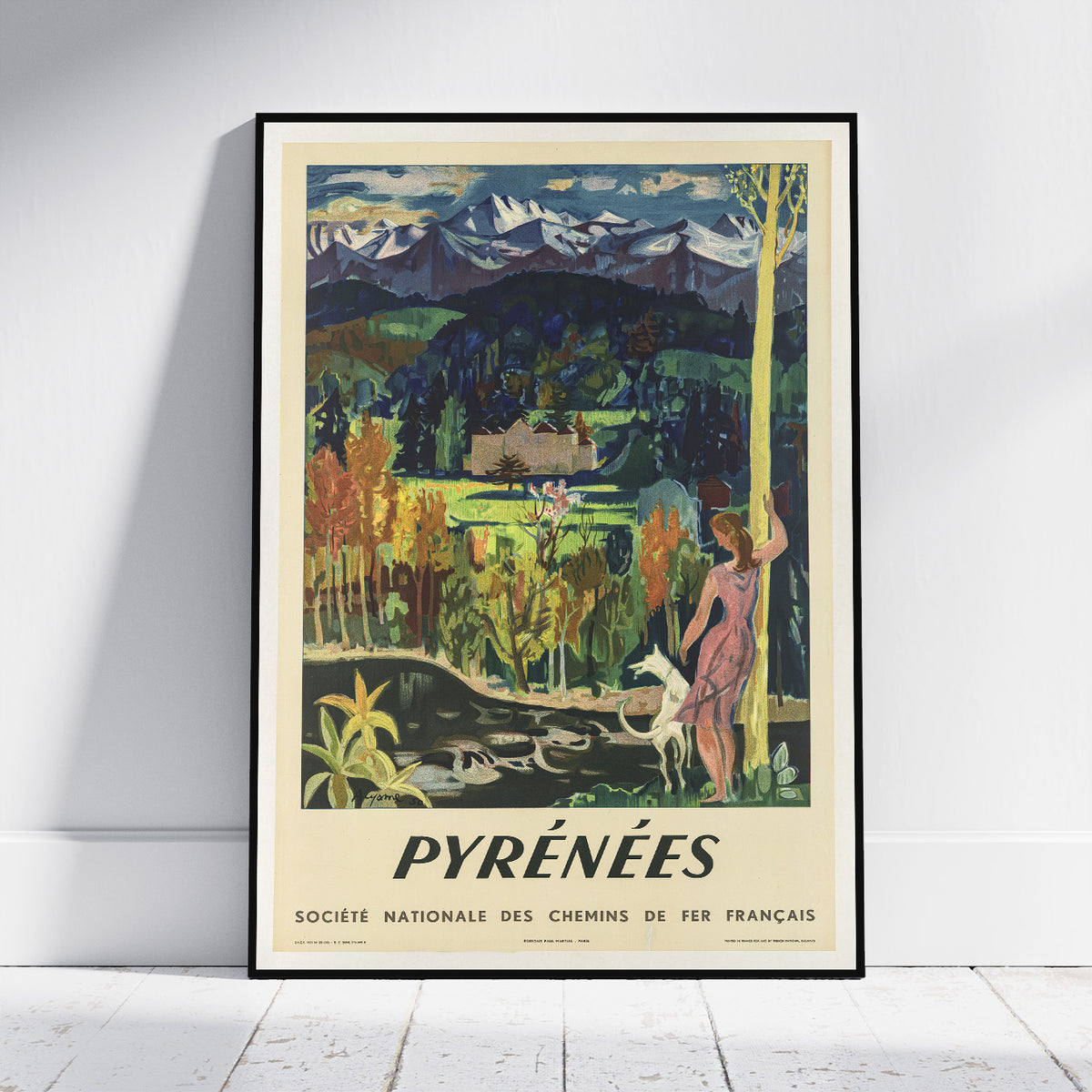 A framed vintage-style travel poster leaning against a white wall on a light wood floor. The poster depicts a scenic mountain landscape with the text 'PYRÉNÉES' and smaller French text below, plus 'Société Nationale des Chemins de Fer Français' at the bottom.