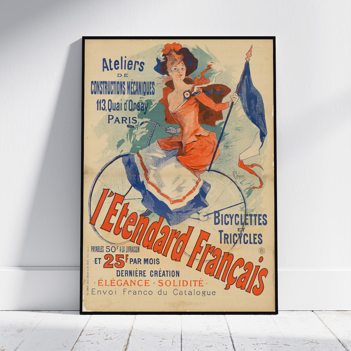 A framed vintage-style poster leaning against a white wall on a light wood floor. The poster features an illustrated woman in a red dress riding a bicycle with large red and blue text reading “L’Etendard Français” and “Bicycles et Tricycles.