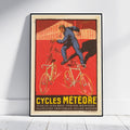 A framed vintage-style poster leaning against a white wall on a light wood floor. The poster artwork shows a cyclist in blue riding bicycles against a bold red background, with the text 'CYCLES MÉTÉORE' and smaller French text at the bottom.