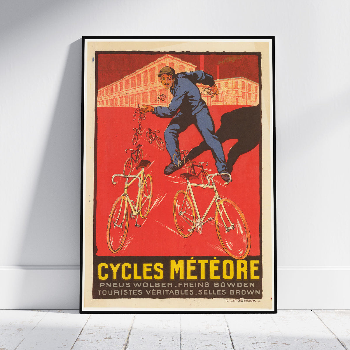 A framed vintage-style poster leaning against a white wall on a light wood floor. The poster artwork shows a cyclist in blue riding bicycles against a bold red background, with the text 'CYCLES MÉTÉORE' and smaller French text at the bottom.