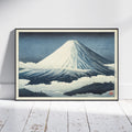 A framed Japanese-style art print of Mount Fuji leaning against a white wall on a white wooden floor. The print features a snow-covered mountain under a dark blue sky, with a black frame and a light beige mat.
