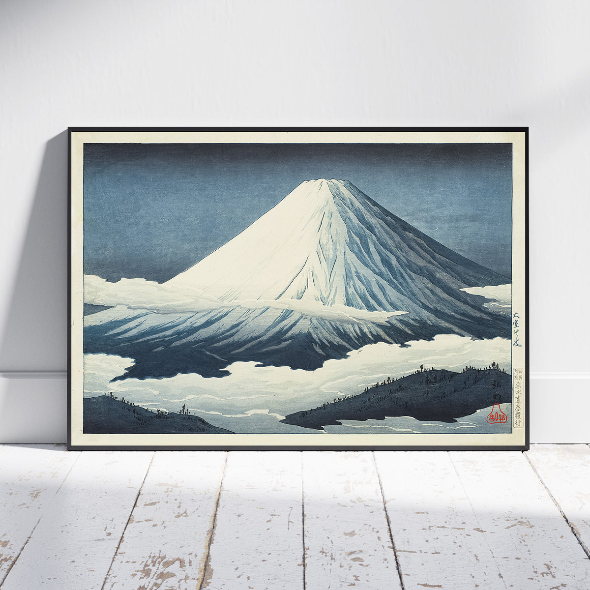 A framed Japanese-style art print of Mount Fuji leaning against a white wall on a white wooden floor. The print features a snow-covered mountain under a dark blue sky, with a black frame and a light beige mat.