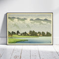 A framed landscape art print leaning against a white wall on a white wooden floor. The print depicts a serene lakeside village with green fields, trees, small houses, distant mountains, and a cloudy sky.