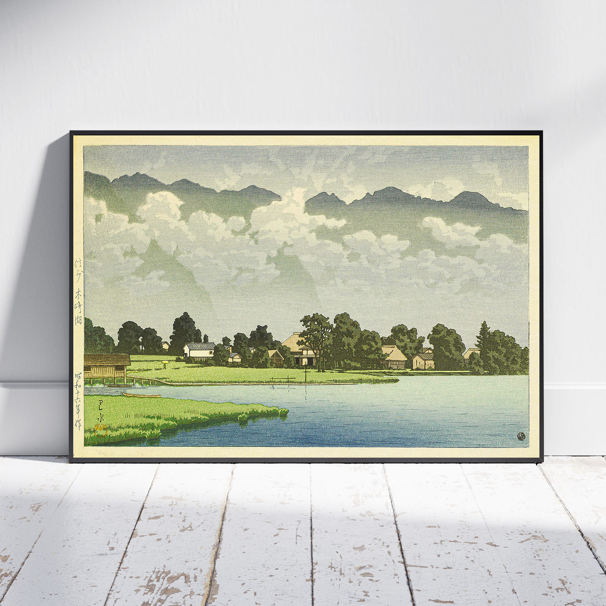 A framed landscape art print leaning against a white wall on a white wooden floor. The print depicts a serene lakeside village with green fields, trees, small houses, distant mountains, and a cloudy sky.