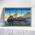 A framed Japanese-style art print depicting three large wooden ships in blue water under a crescent moon, leaning against a white wall on a light wood floor.