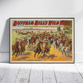 A framed vintage Western poster leaning against a white wall on a white wood floor. The poster reads 'Buffalo Bill's Wild West' and shows a crowded scene of riders on horseback in a desert landscape.