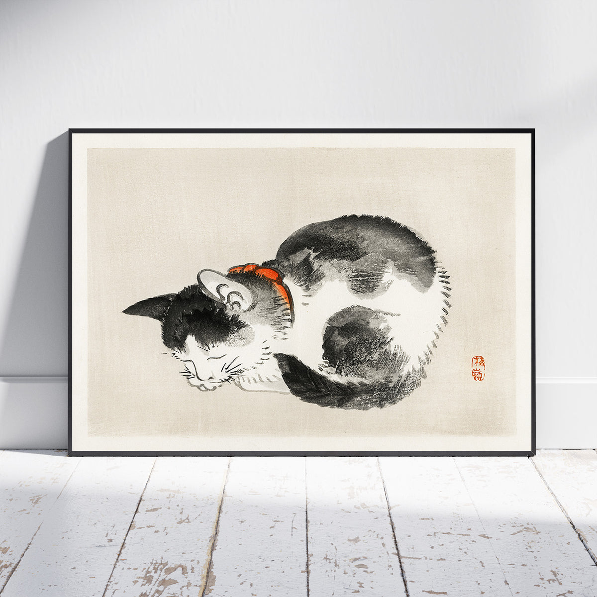 A framed art print featuring a black and white cat curled up in a sleeping pose with a small orange collar, displayed against a white wall on a light wood floor.