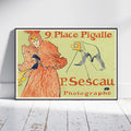 A framed vintage poster leaning against a white wall on a white wood floor. The poster features an Art Nouveau illustration of a woman in a flowing orange dress with a camera and the text '9.