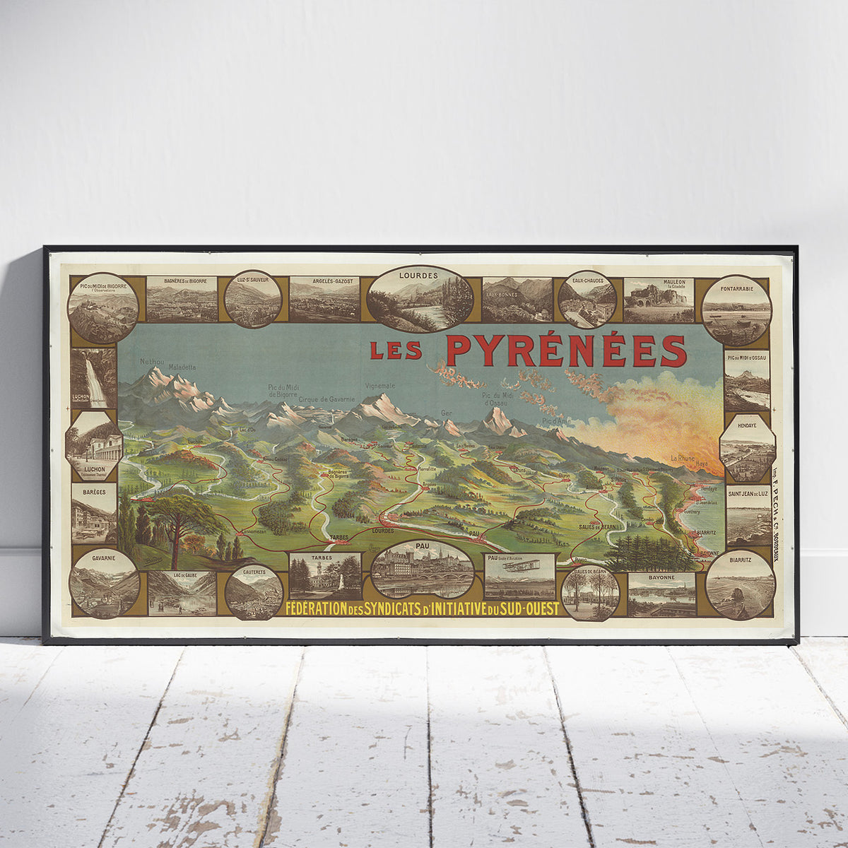 A framed vintage-style poster reading “LES PYRÉNÉES” leaning against a white wall on a white wood floor. The artwork shows a mountain landscape with roads, trees, and small inset scenic panels around the border.